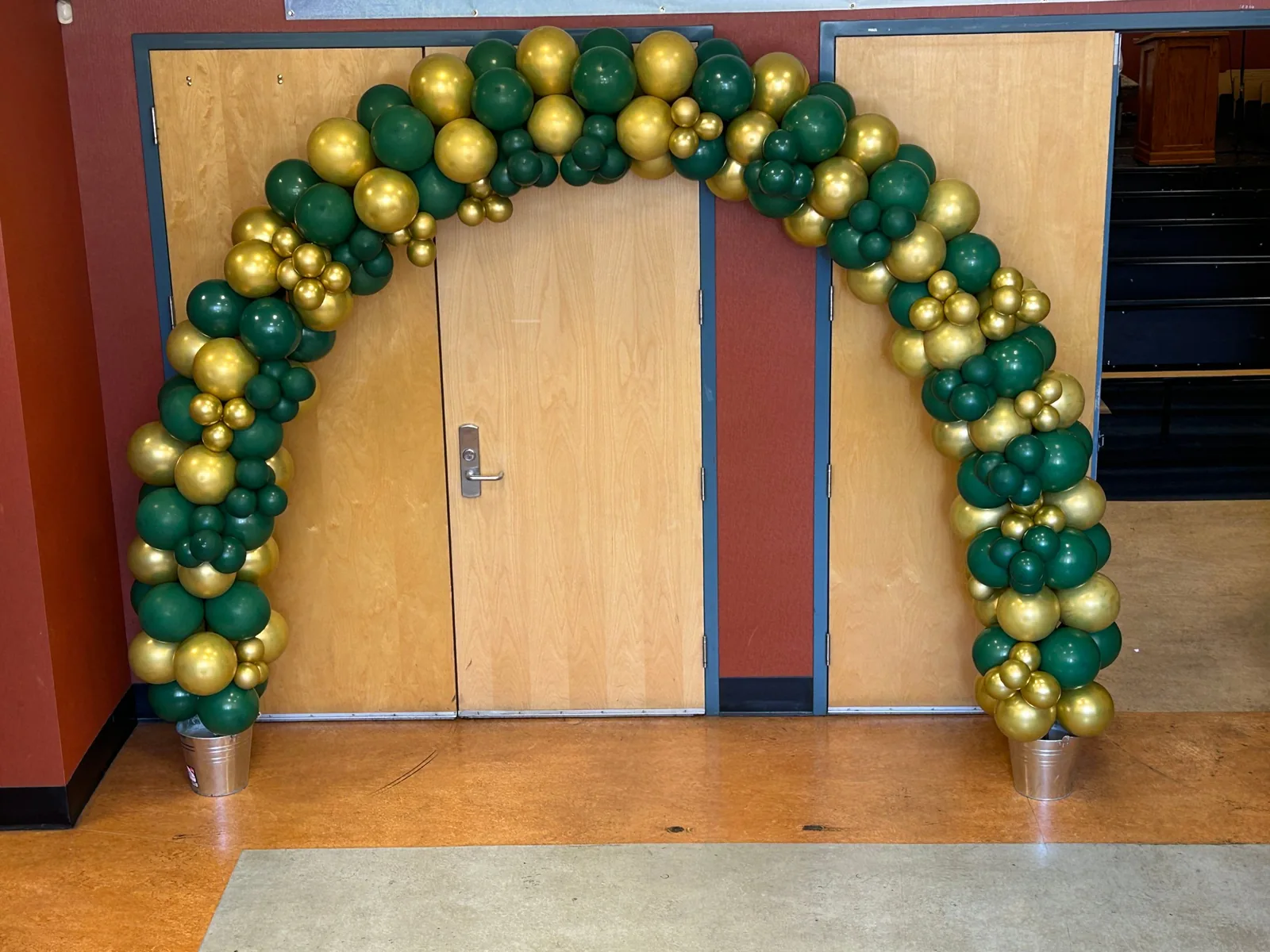 Forest green and chrome gold organic balloon arch framing a set of double doors at an indoor entrance
