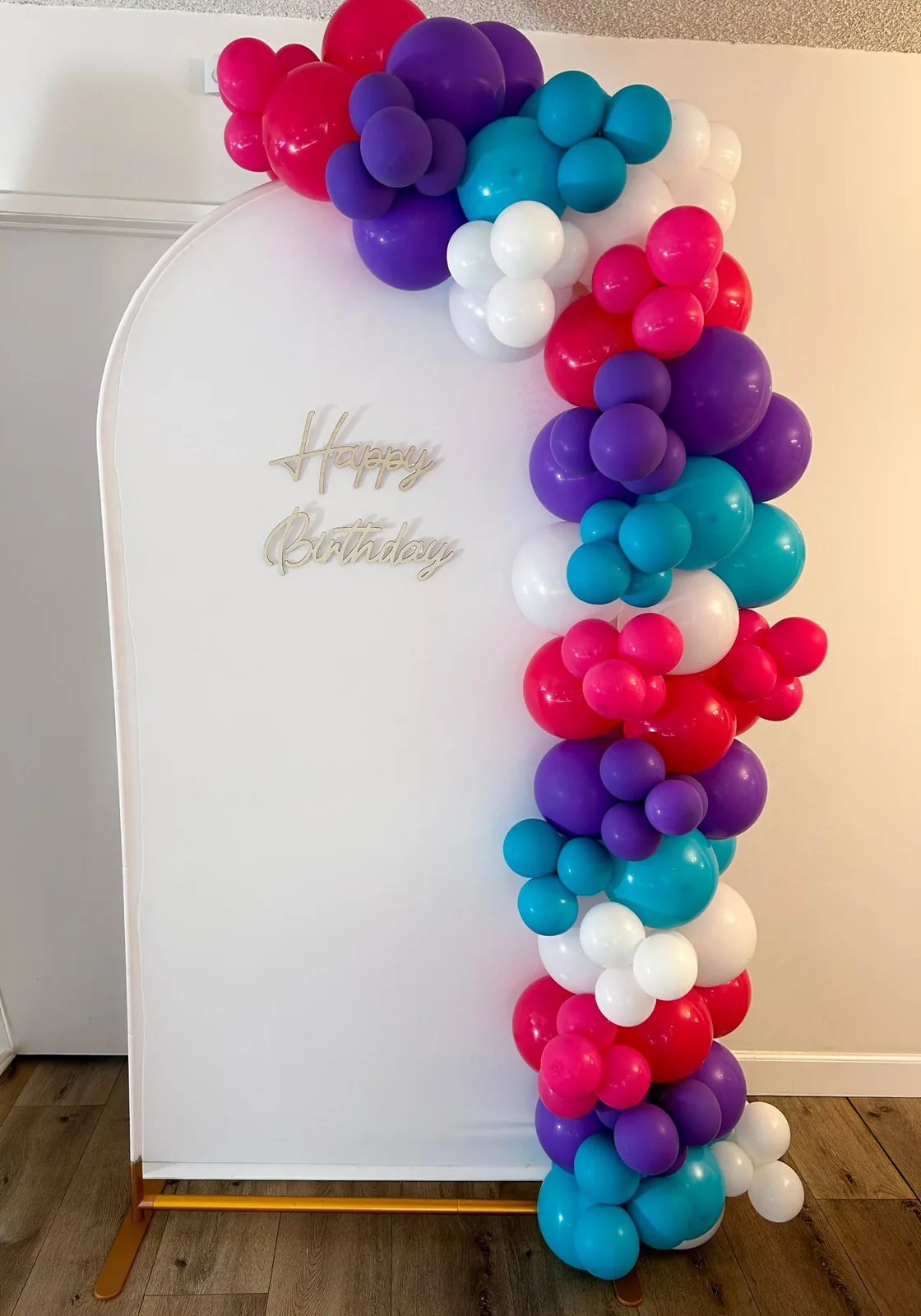 Pink, purple, teal and white balloon garland on a white chiara arch backdrop with a gold Happy Birthday sign